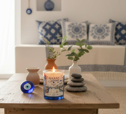 Lit Aegean Shores coconut wax candle by Rene's Scented Spark displayed on a wooden coffee table with stacked spa stones, greenery, and blue Mediterranean décor in a peaceful living room.