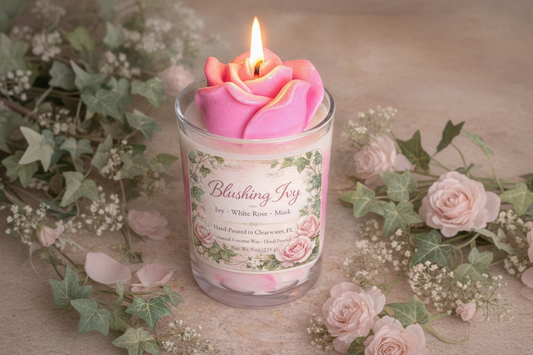 Lit Blushing Ivy candle with sculpted pink rose wax top in a glass jar, featuring ivy and rose label, styled with flowers and soft greenery