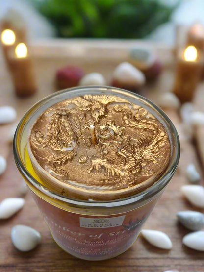 Spiritual candle collection displayed on a wooden table with Ember of Strength candle topped with phoenix and dragon, and Tranquil Spirit candle beside a Buddha figure. Surrounded by crystals, stones, and greenery — symbolic gift candles for meditation, healing, and renewal