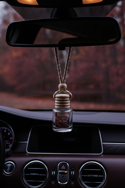 Haunted Orchid hanging car air freshener suspended from a rearview mirror inside a dark, elegant car interior, with moody autumn scenery visible through the windshield.