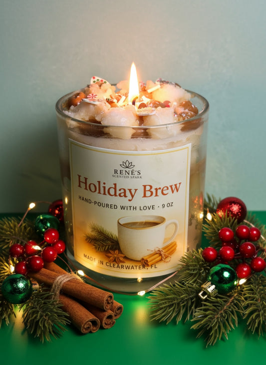 Lit candle in a glass jar labeled 'Holiday Brew' with festive decorations on a green surface.