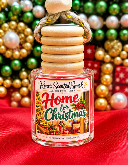 A clear glass car air freshener bottle with a stacked light wood cap and rope handle, labeled 'Home for Christmas' by Rene's Scented Spark, set on red fabric with a blurred Christmas ornament background.