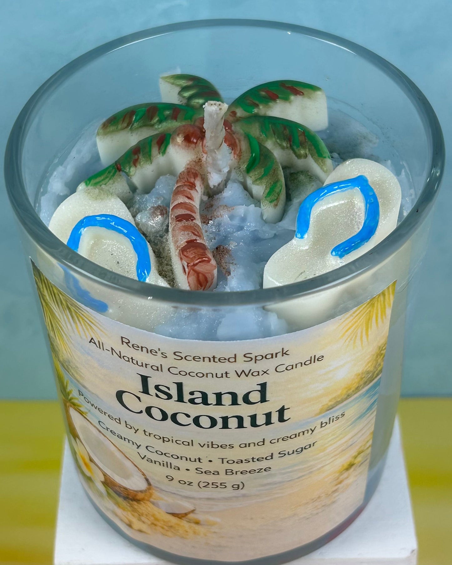 Candle labeled 'Island Coconut' with a label featuring coconut imagery on a light blue background with flip flop molds and a palm tree