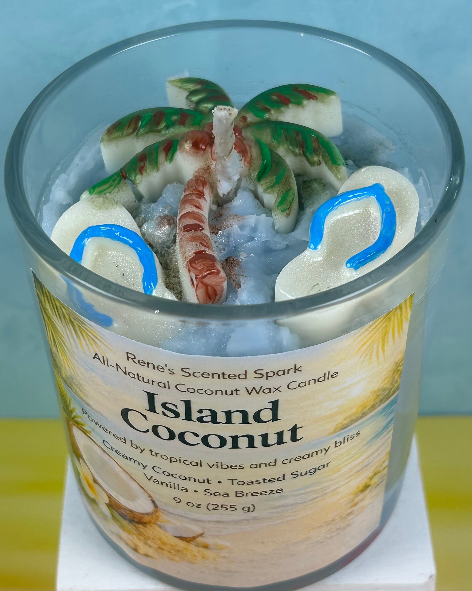 Candle labeled 'Island Coconut' with a label featuring coconut imagery on a light blue background with flip flop molds and a palm tree