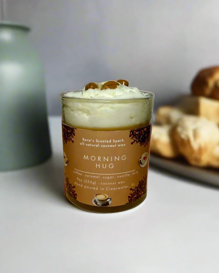 Morning Hug candle glowing beside a steaming coffee mug, books, and cozy blanket — coffee and caramel candle perfect for gifting