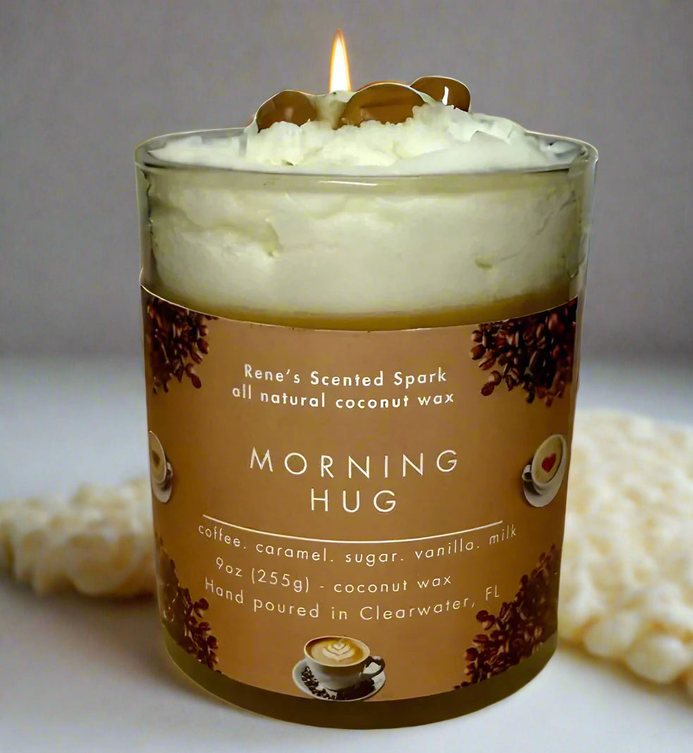 Morning Hug candle glowing beside a steaming coffee mug, books, and cozy blanket — coffee and caramel candle perfect for gifting.