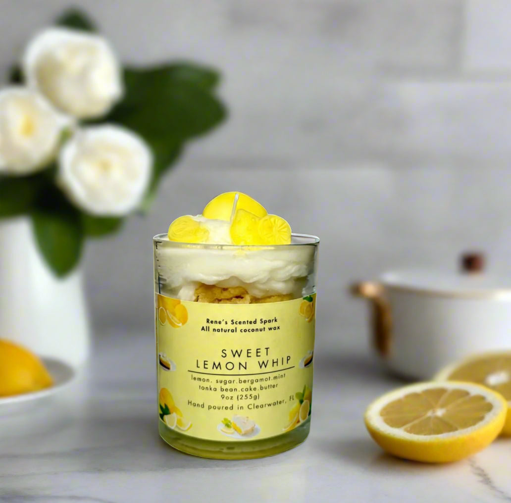 Sweet Lemon Whip candle glowing on a kitchen counter beside lemons, a slice of cake, and fresh flowers — citrus dessert candle perfect for gifting.