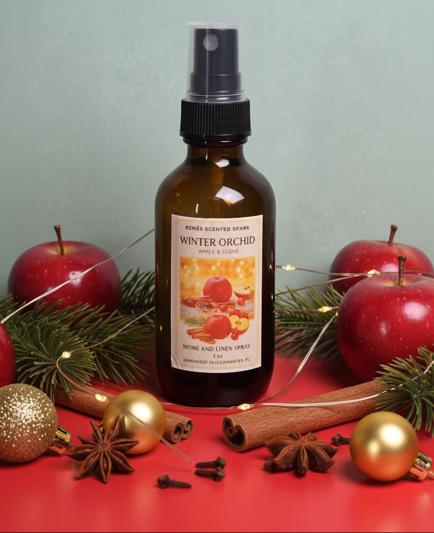Bottle of 'Winter Orchard' home and linen spray surrounded by apples, cinnamon sticks, and Christmas ornaments on a red surface.