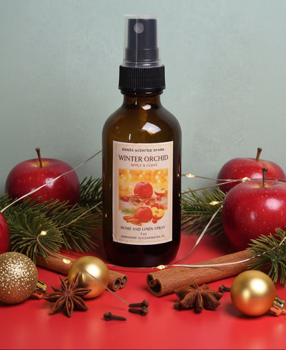 Bottle of 'Winter Orchard' home and linen spray surrounded by apples, cinnamon sticks, and Christmas ornaments on a red surface.