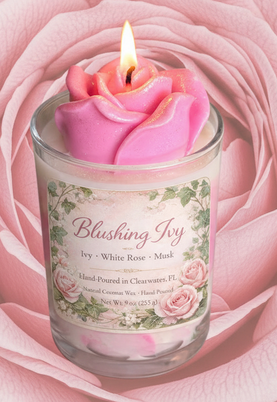 Lit Blushing Ivy candle with sculpted pink rose wax top in a glass jar, featuring floral label and set against a large soft pink rose backdrop