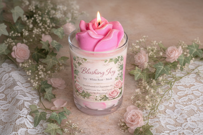 Lit Blushing Ivy candle with sculpted pink rose wax top in a glass jar, styled with lace, ivy greenery, and delicate pink roses