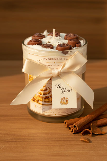 A 9 oz clear glass cinnamon roll candle jar with a warm label sits styled as a gift on a warm wood surface. A cream ribbon, cinnamon sticks, and a small gift tag are placed beside it. Soft warm natural light. 