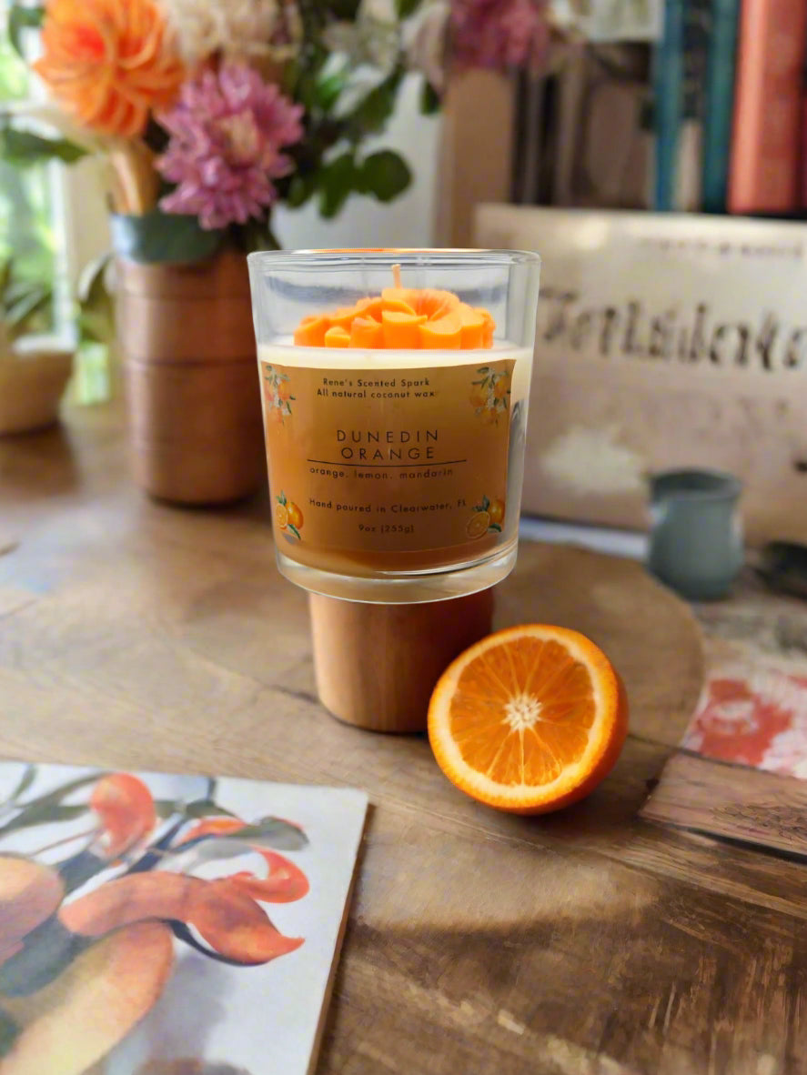 Dunedin Orange candle glowing on a table with fresh oranges and flowers, creating a cozy citrus-inspired atmosphere, ideal as a gift.