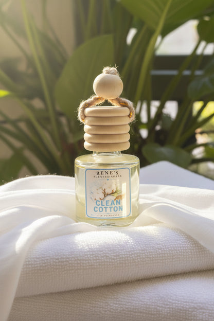 Clean Cotton hanging car air freshener by Rene’s Scented Spark displayed on soft white folded linen in warm sunlight with green tropical leaves in the background.