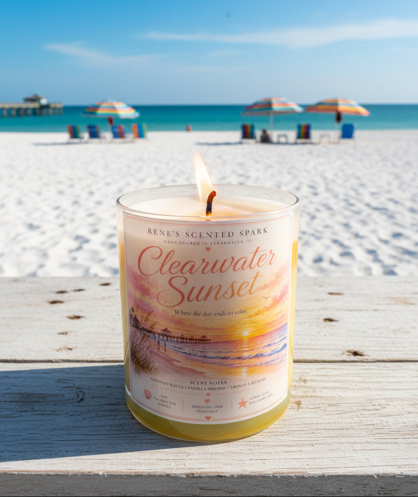 Lit Clearwater Sunset hand-poured coconut wax candle displayed on a wooden table overlooking the white sands of Clearwater Beach. Coastal-inspired candle with colorful beach umbrellas and turquoise Gulf waters in the background.