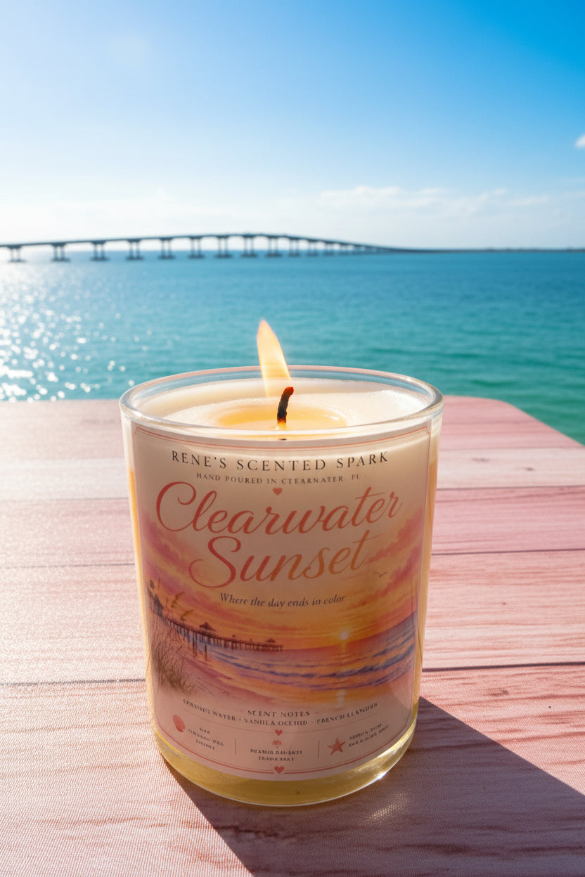 Lit Clearwater Sunset hand-poured coconut wax candle displayed on a waterfront table overlooking turquoise Gulf waters and a scenic bridge. Coastal-inspired candle celebrating the beauty of Clearwater, Florida.