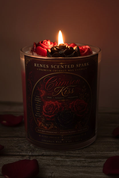 Crimson Kiss coconut wax candle glowing in a dark romantic setting with crimson and black rose wax toppers and scattered red rose petals.