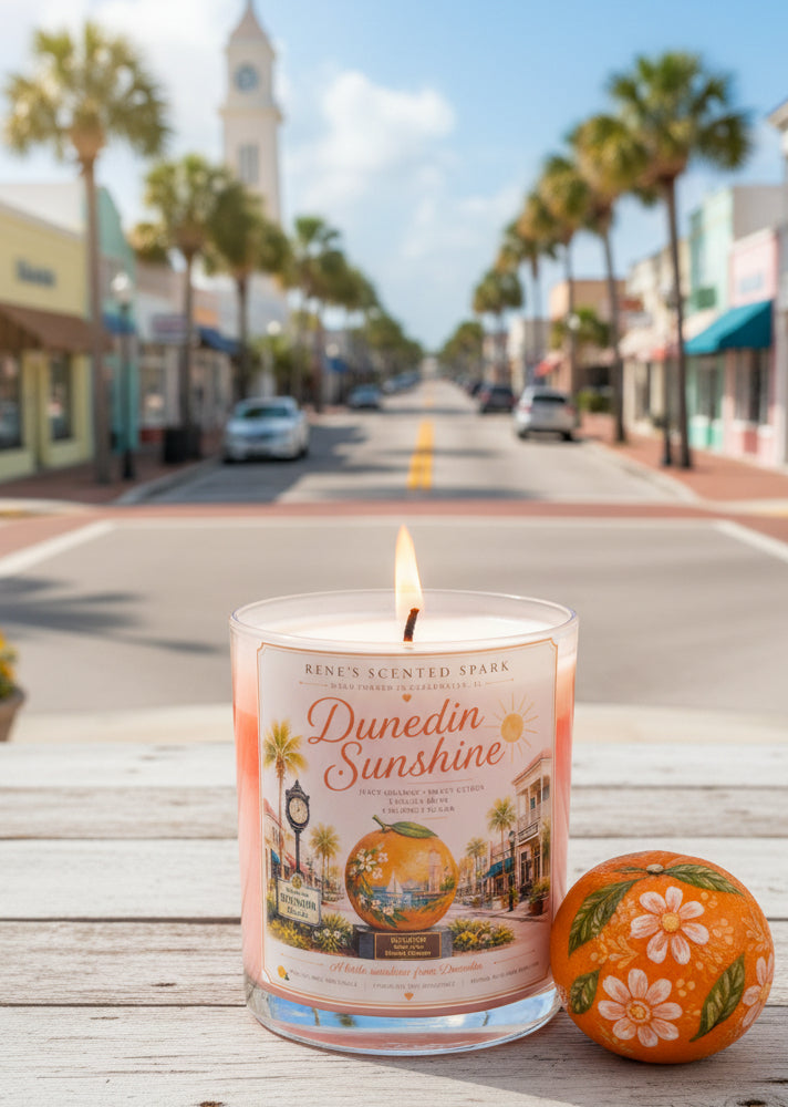 Lit Dunedin Sunshine hand-poured coconut wax candle displayed in historic downtown Dunedin, Florida. Coastal-inspired candle featuring citrus artwork with an orange and palm-lined Main Street in the background.