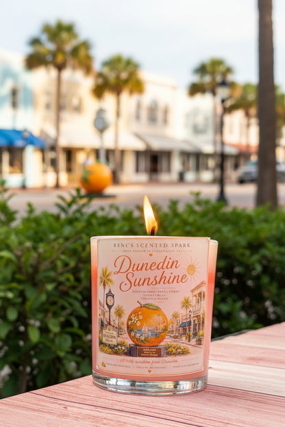 A lit scented candle in a clear glass jar sits on a wooden surface outdoors, with a blurred street scene featuring palm trees and buildings in the background. The label reads 'Dunedin Sunshine' Renes Scented Spark.