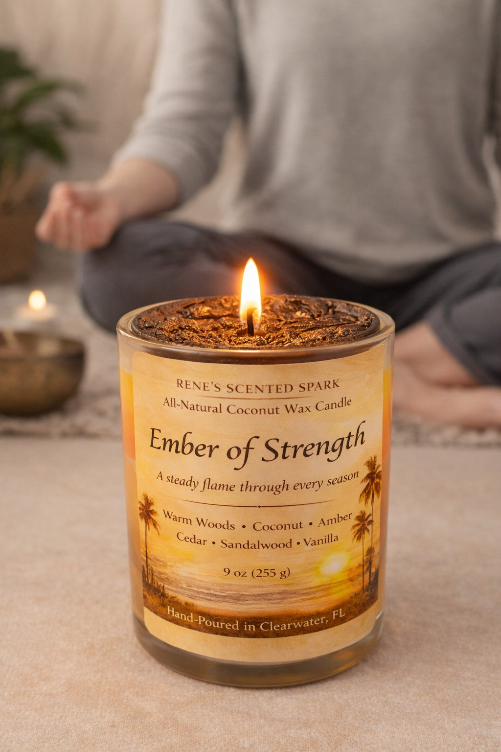Ember of Strength” coconut wax candle in a clear glass jar, featuring a golden sunset island label with palm trees and warm amber tones. The candle glows softly against a calm, neutral meditation-inspired background, conveying grounding, resilience, and quiet strength