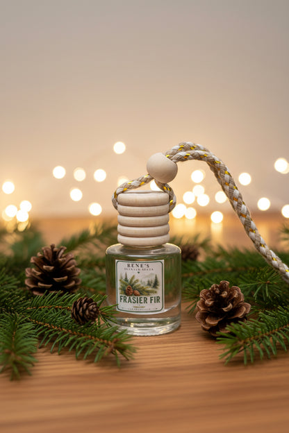 Frasier Fir car air freshener with wooden diffuser cap surrounded by evergreen branches and pinecones, with warm holiday lights softly glowing in the background. 🌲✨