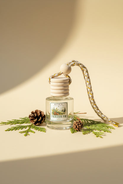 Frasier Fir car air freshener with wooden diffuser cap styled with evergreen sprigs and pinecones on a warm cream background with soft sunlight. 🌲✨
