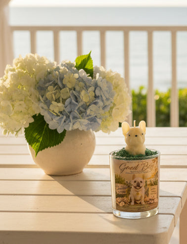 A luxury candle featuring a handcrafted french bull  dog wax topper displayed on a white coastal porch overlooking the ocean. Soft sea breezes move sheer curtains while fresh flowers decorate the table.