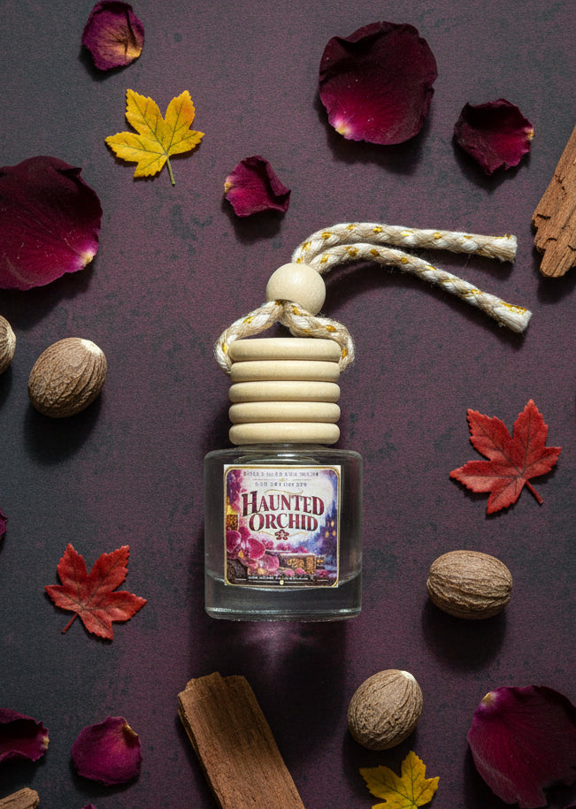 Haunted Orchid hanging car air freshener styled on a dark burgundy background with deep orchid petals, autumn leaves, warm wood pieces, and nutmeg for a moody fall fragrance display.