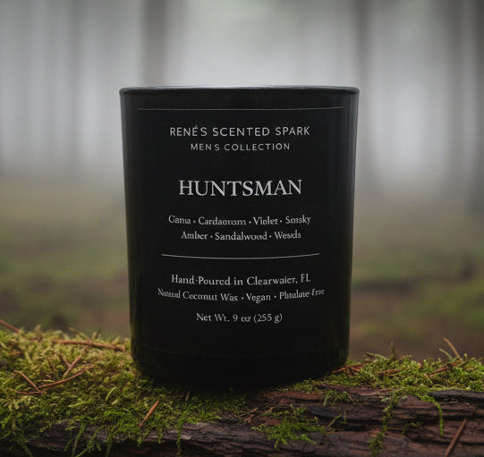 Black luxury candle labeled “Huntsman” by Rene’s Scented Spark placed on moss and bark in a foggy forest setting