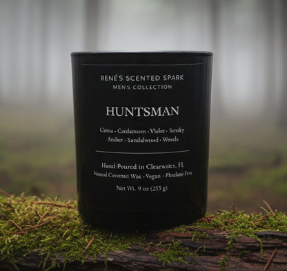 Black luxury candle labeled “Huntsman” by Rene’s Scented Spark placed on moss and bark in a foggy forest setting
