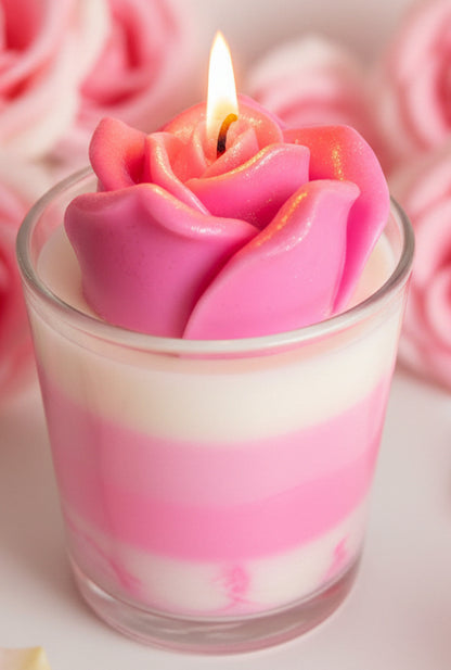 Close-up of a lit pink rose-shaped candle with layered pink and white wax in a glass jar, set against a soft floral background