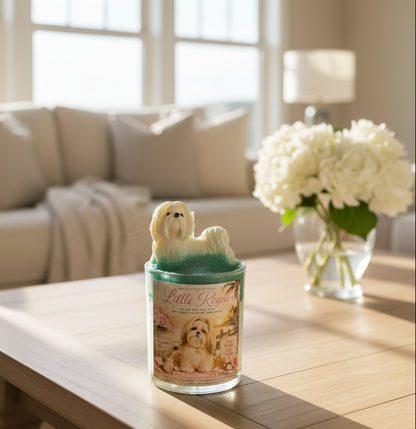 A hand-poured luxury candle with a detailed white Maltese dog wax topper displayed on a light wood coffee table in an elegant coastal living room. Soft morning sunlight streams through large windows, with cream furnishings, fresh flowers, and a cozy throw blanket in the background.