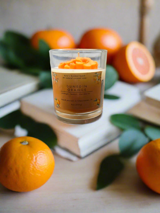 Dunedin Orange candle glowing on a wooden table with fresh oranges, in a cozy sunlit room
