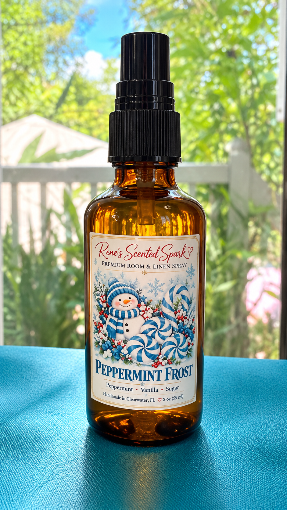 Rene's Scented Spark Peppermint Frost 2 oz room spray in amber glass bottle with blue snowman label on teal fabric outdoors
