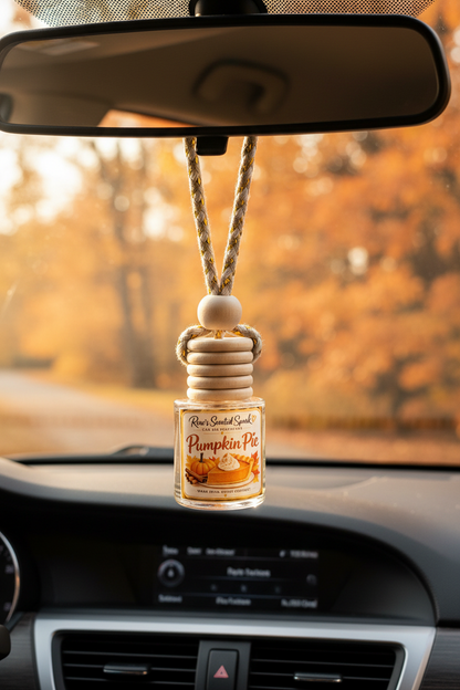 Pumpkin Pie car freshener by Rene’s Scented Spark hanging from a rearview mirror with warm golden autumn foliage visible through the windshield.