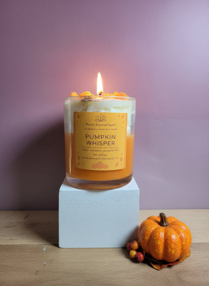 Candle labeled 'Pumpkin Whisper' with a small pumpkin on a wooden surface against a purple background