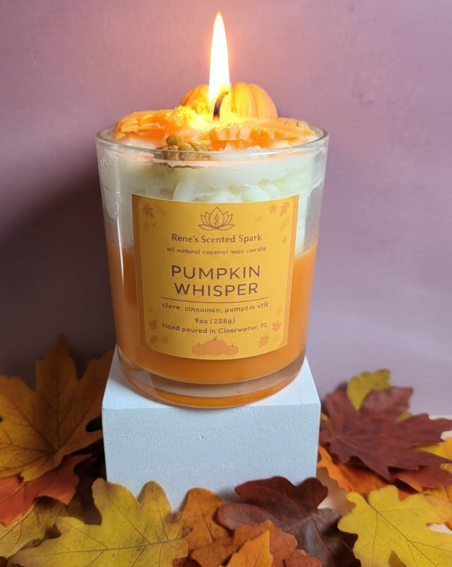 Candle labeled 'Pumpkin Whisper' surrounded by autumn leaves on a light purple background