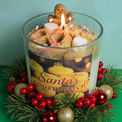 Decorative candle with gingerbread man and cookies, surrounded by Christmas ornaments and greenery.