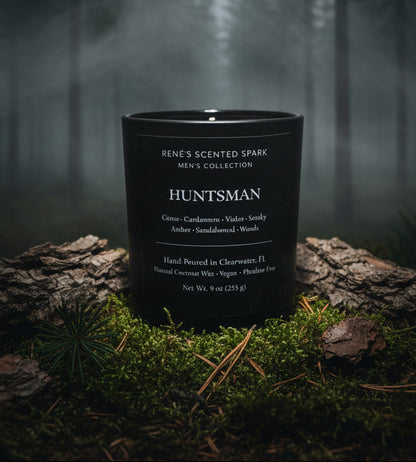 Black candle labeled 'Huntsman' from Rene's Scented Spark Men's Collection on a forest floor.