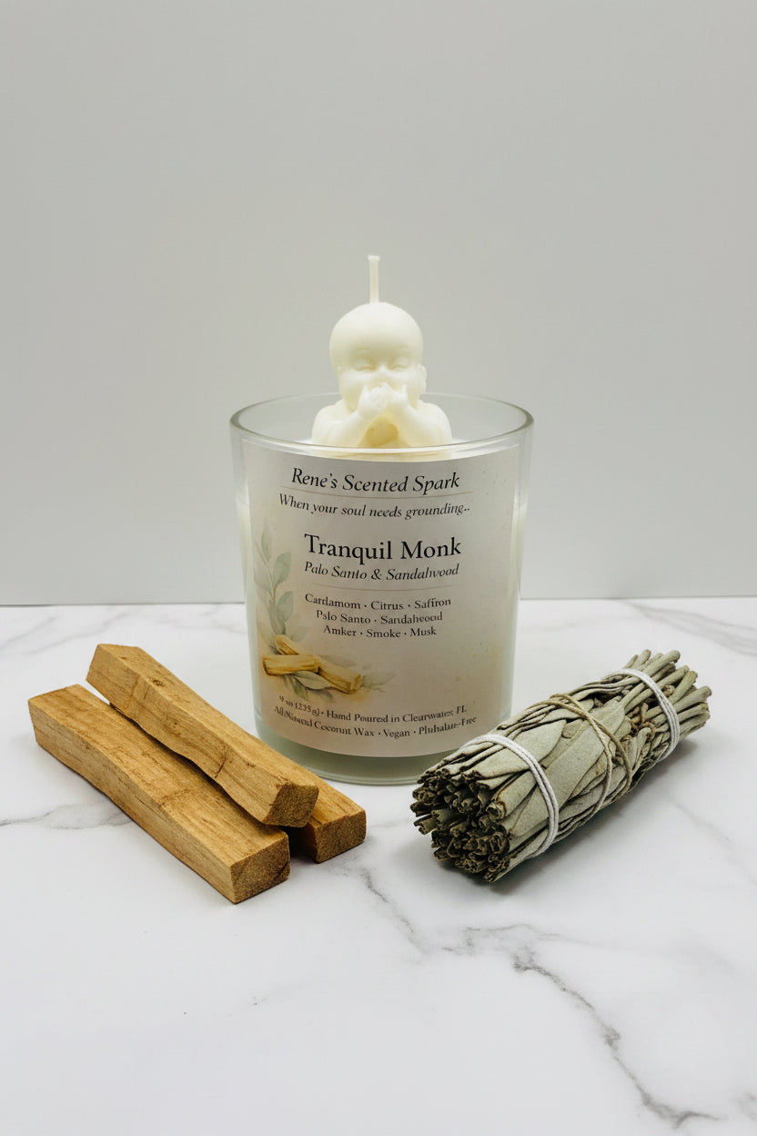 Tranquil Monk coconut wax candle with a sculpted meditating monk topper, displayed with palo santo wood and a sage bundle on a white marble surface.