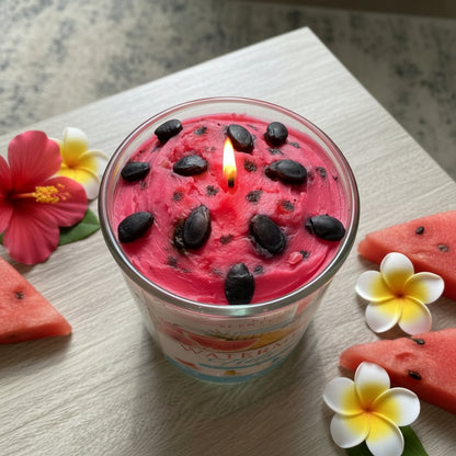 Watermelon Tide Candle 9 oz clear glass candle jar showing a lush pink whipped wax top with realistic dark oval watermelon seed mold accents in sharp detail with tropical flowers and watermelon slices
