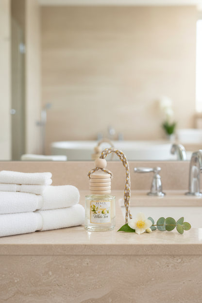 White Tea car freshener in a clear glass bottle displayed on a neutral bathroom vanity beside folded white towels, a white jasmine flower, and eucalyptus greenery.