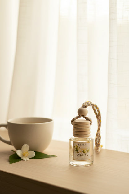 White Tea car freshener in a clear glass bottle beside a warm cup of tea and white jasmine flower, styled on a light wood surface by a softly sunlit window.