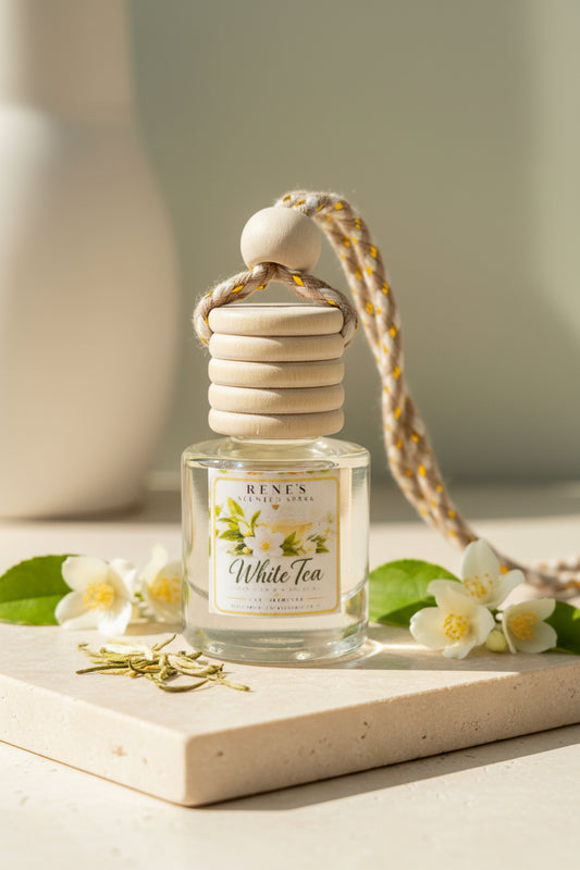 White Tea hanging car freshener in a clear glass bottle with a natural wood cap, styled with white jasmine flowers and green leaves on a cream stone surface in soft sunlight.