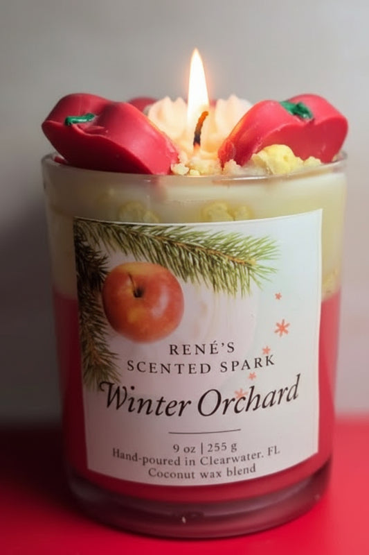 A handcrafted coconut wax candle labeled “Winter Orchard” by René’s Scented Spark. The candle is topped with decorative red apple wax pieces and cream-colored wax swirls, evoking a festive holiday feel. The label features an apple and pine branch design, highlighting the cozy Christmas theme.