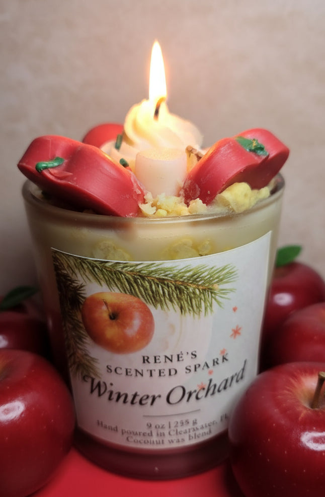A handcrafted coconut wax candle labeled “Winter Orchard” by René’s Scented Spark. The candle is topped with decorative red apple wax pieces and cream-colored wax swirls, evoking a festive holiday feel. The label features an apple and pine branch design, highlighting the cozy Christmas theme.