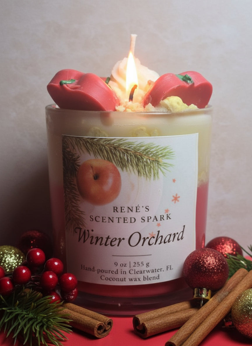 A handcrafted coconut wax candle labeled “Winter Orchard” by René’s Scented Spark. The candle is topped with decorative red apple wax pieces and cream-colored wax swirls, evoking a festive holiday feel. The label features an apple and pine branch design, highlighting the cozy Christmas theme.