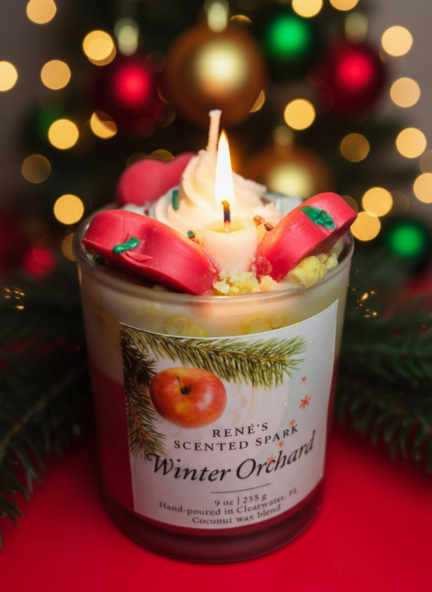 A handcrafted coconut wax candle labeled “Winter Orchard” by René’s Scented Spark. The candle is topped with decorative red apple wax pieces and cream-colored wax swirls, evoking a festive holiday feel. The label features an apple and pine branch design, highlighting the cozy Christmas theme.